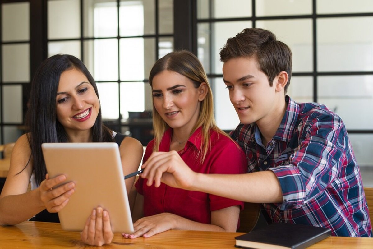 Three college students sit at a table, smiling and engaged, as they explore the best study apps for college students on a tablet in their office setting.