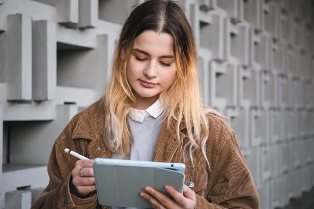 A student in a brown jacket is engrossed in using a tablet with a stylus, perhaps exploring the best study apps for college students, while standing before a textured wall.